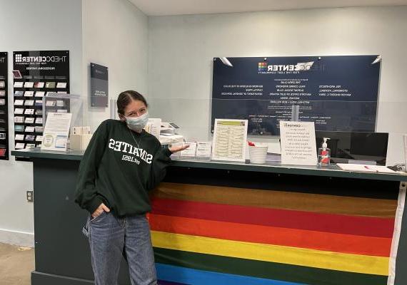 Elana standing in front of a desk with a Rainbow flag