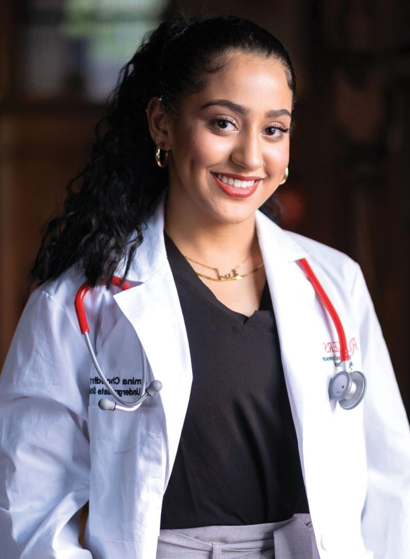 Amina Choudhry smiling, wearing a white lab coat and stethoscope