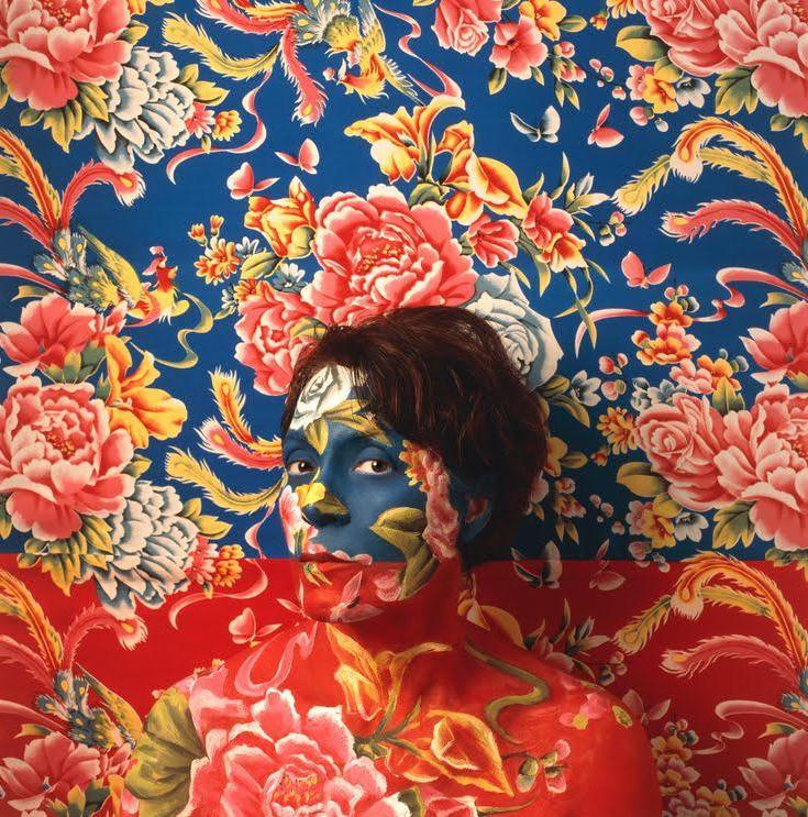 艺术家的自画像戴着蓝色和红色的花妆作为伪装，背后是壁纸